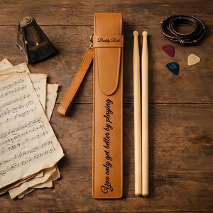 Puede incluir: Un estuche de baquetas de cuero marrón con el texto "You only get better by playing" y "Buddy Rich". Dos baquetas de madera, púas de guitarra y una pulsera trenzada sobre una superficie de madera.