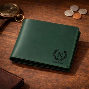 Puede incluir: Una cartera de cuero verde oscuro con un diseño personalizado. La cartera presenta una corona de laurel negra con la letra "N" y el nombre "Nathan" grabado en la parte delantera. La cartera está sobre una superficie de madera con una brújula y monedas.