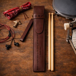 Può includere: Un astuccio per bacchette in pelle marrone con le iniziali "G.M.P" e il testo "Make a joyful noise to the Lord, all the earth! - Psalm 98:4". Due bacchette di legno sono accanto all'astuccio. Sono visibili anche una bandana rossa, una croce dorata e auricolari neri.