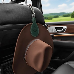 Puede incluir: Un sombrero de vaquero marrón colgado del reposacabezas de un coche. El sombrero se sujeta con una correa de cuero. Una etiqueta de cuero verde con un emblema de ancla está sujeta a un mosquetón plateado.