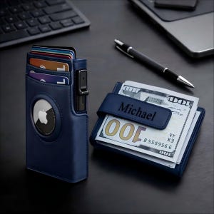 Pode incluir: Conjunto de carteira e porta-cartões em couro azul marinho. O porta-cartões possui um Apple AirTag e comporta vários cartões. A carteira, com o nome "Michael" na alça, está cheia de notas de 100 dólares americanos. Uma caneta preta está por perto.