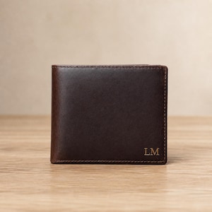 May include: A dark brown leather bifold wallet featuring visible stitching along the edges and the embossed capital letters LM in the bottom right corner. The wallet has a smooth, matte finish and a rectangular shape with rounded corners.