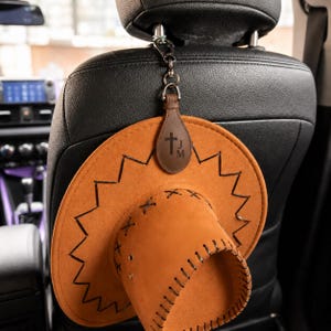 May include: A tan cowboy hat with a zig-zag brim design hangs from a car headrest. The hat is attached to a leather tag with a cross and initials. The hat is made of a soft material and has decorative stitching.