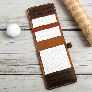May include: A brown leather golf score card holder with the text "The harder you work, the luckier you get." and "Golf is a game of inches the most important ones are between your ears." A white golf ball is next to the holder.