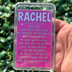 以下が含まれることがあります： ピンク色のインサートが入った透明なプラスチック製の医療用IDバッジ。「RACHEL medical assistant」と祈りの言葉が書かれています。バッジは緑の葉の背景に対して保持されています。