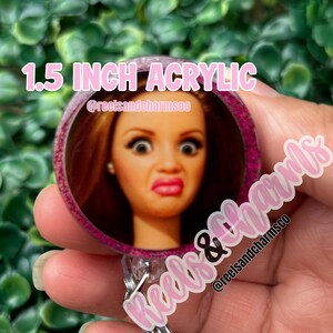 May include: A 1.5-inch acrylic badge reel featuring a doll's face with a surprised expression. The badge reel has a pink glitter border and the text "1.5 INCH ACRYLIC" in pink. The @reelsandcharmsco logo is also visible.