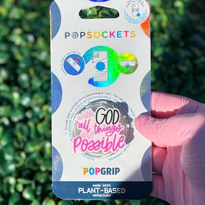 May include: A PopSockets PopGrip with the phrase "With God All Things Are Possible" in pink and black lettering. The packaging is white and blue with holographic accents and the PopSockets logo.