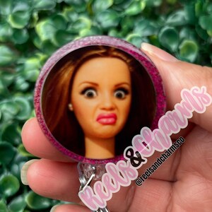 May include: A round, pink and purple badge reel featuring a doll's face with a surprised expression. The badge reel has a glittery border and the text "Reels & Charms" in a cursive font. The @reelsandcharmsco is also visible.
