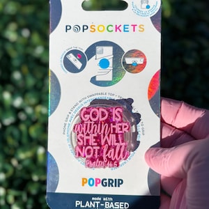 May include: A pink PopSocket phone grip with the text "GOD IS within HER SHE WILL NOT fall Psalm 46:5". The PopSocket is on a white and iridescent background. The packaging also includes instructions and the PopSocket brand name.