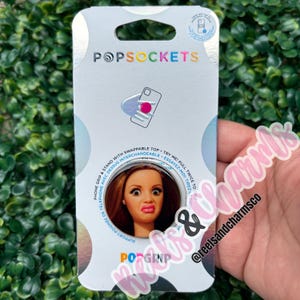 May include: A PopSockets PopGrip phone stand and grip in its original retail packaging. The white card features the PopSockets logo, a diagram of the product, and a circular cutout displaying a graphic of a person with a surprised expression.