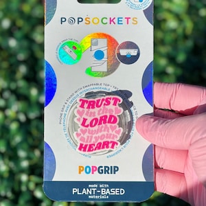 Puede incluir: Un PopSockets PopGrip con fondo blanco y las palabras "Trust in the Lord with all your heart" en rosa. El PopGrip está en una tarjeta con el logotipo de PopSockets y las palabras "Plant-Based materials".