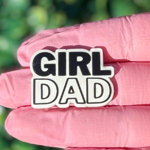 Puede incluir: Un dije blanco y negro con las palabras "GIRL DAD" en una fuente en negrita y en bloque. El dije es sostenido por una persona que usa guantes rosas, con un fondo verde borroso.
