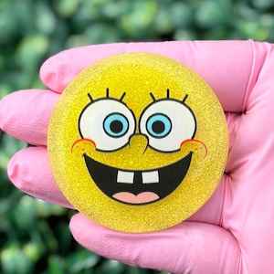 May include: A yellow, glittery, circular item featuring a cartoon face with large eyes, a wide smile, and two teeth. The character's face is yellow with black outlines and blue eyes. The item is held by a person wearing pink gloves.