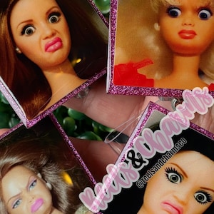 Puede incluir: Primer plano de cuatro insignias rectangulares con caras de muñecas con expresiones exageradas. Las insignias tienen bordes de purpurina rosa y se mantienen juntas. El texto "Reels & Charms" es visible en una insignia.