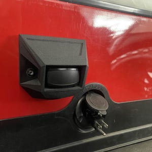 May include: A black, angular charging port cover and a charging port are mounted on a red surface. The charging port has a black cover with the word "CHARGER" visible. The charging port has a plug inserted.