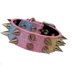 May include: A pastel rainbow glitter collar featuring two rows of sharp, silver-toned metal spikes. The collar includes a silver-toned metal buckle and eyelets for adjustment. The text "DAUNTING CLAWS" is visible in the bottom left corner.