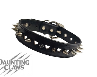 May include: Black leather collar with silver-colored spikes and heart-shaped cutouts. The collar has a buckle closure and is designed with a punk rock aesthetic. The brand name "Daunting Claws" is visible.