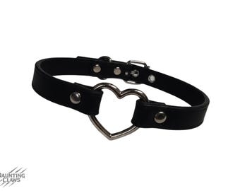 May include: Black choker necklace with a silver heart-shaped pendant. The choker is made of black leather-like material and features a buckle closure. The heart pendant is centered on the choker.