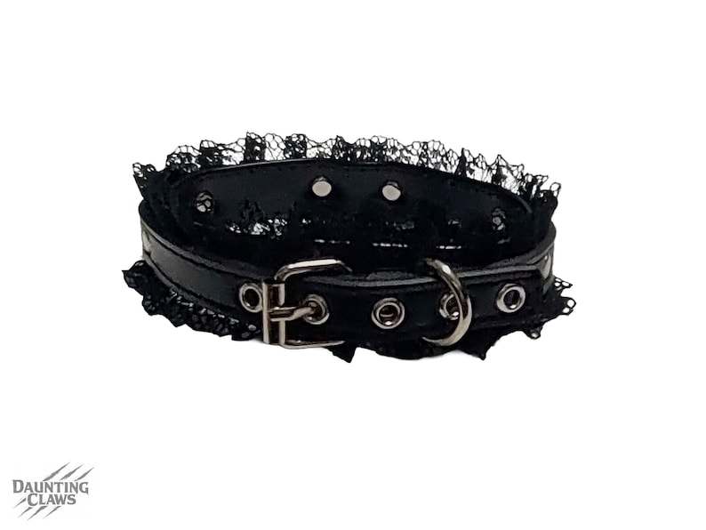 May include: Black choker collar with a lace trim. The collar is made of black leather and features a silver buckle and a metal ring. The brand name "DAUNTING CLAWS" is visible in the bottom left corner.