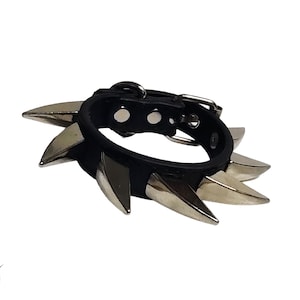 May include: Black leather wristband with silver-colored spikes. The wristband has a buckle closure and is designed with multiple metal spikes around the band. The product is set against a white background.