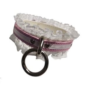 May include: A white lace collar with a pink and silver band and a large silver ring. The collar has a delicate, feminine design, with a soft, romantic aesthetic. The collar is designed to be worn around the neck.