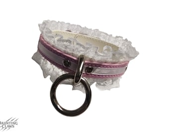 May include: A white lace collar with a pink and silver band and a large silver ring. The collar has a delicate, feminine design, with a soft, romantic aesthetic. The collar is designed to be worn around the neck.