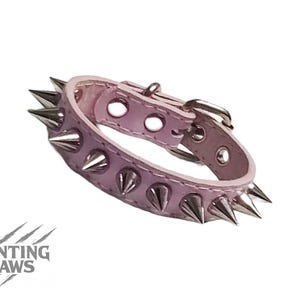 May include: A pale purple leather collar featuring silver-coloured spikes. The collar has a silver buckle and adjustment holes. The brand name "DAUNTING CLAWS" is printed in the lower left corner.