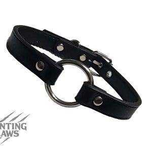Könnte beinhalten: Schwarzes Lederhalsband mit großem silberfarbenem Metallring und Schnallenverschluss. Das Halsband hat silberne Nieten und ist geschwungen geformt. Das Daunting Claws Logo ist unten links sichtbar.