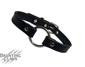 May include: Black leather collar with a large silver metal ring and buckle closure. The collar has silver rivets and is designed with a curved shape. The Daunting Claws logo is visible in the lower left corner.