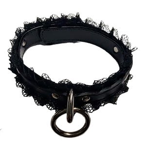 May include: Black choker collar with a lace trim and a silver metal ring. The collar is made of black leather-like material and has silver rivets. The brand name "DAUNTING CLAWS" is visible in the lower left corner.