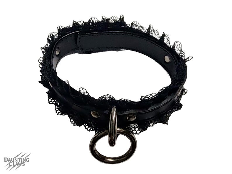 May include: Black choker collar with a lace trim and a silver metal ring. The collar is made of black leather-like material and has silver rivets. The brand name "DAUNTING CLAWS" is visible in the lower left corner.