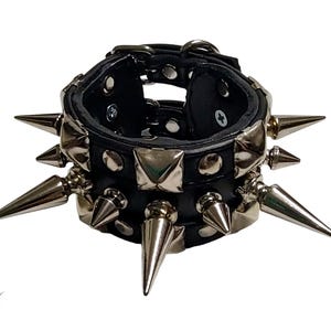 Op de afbeelding: Zwarte leren armband met zilverkleurige spikes en vierkante studs. De armband heeft een gespsluiting en is ontworpen met meerdere rijen scherpe, puntige metalen studs. Een opvallend accessoire.