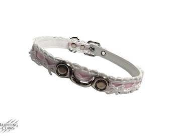 May include: A decorative cat collar in white and pink, featuring a lace design. The collar has silver-coloured metal hardware, including a buckle and D-rings. The collar is made of a white leather-like material and pink fabric.