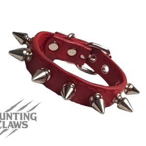 May include: Red leather collar with silver cone studs and a buckle fastening. This striking collar is designed to make a statement. The Daunting Claws logo is visible. The collar is a bold accessory.