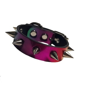 May include: A multi-colored, iridescent leather bracelet with silver-colored metal spikes. The bracelet has a buckle closure and is primarily shades of pink, purple, and green. The brand name "DAUNTING CLAWS" is visible.