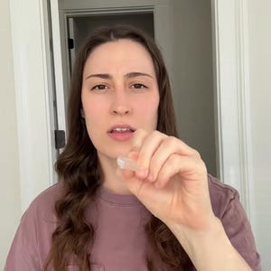 May include: A clear, small, crystal-like object is held up in front of a person. The person is wearing a mauve-colored t-shirt. The background is a white door frame and wall.