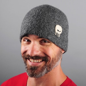 May include: A gray wool beanie with a felt skull and crossbones design. The beanie is worn by a person with a beard and a smile.