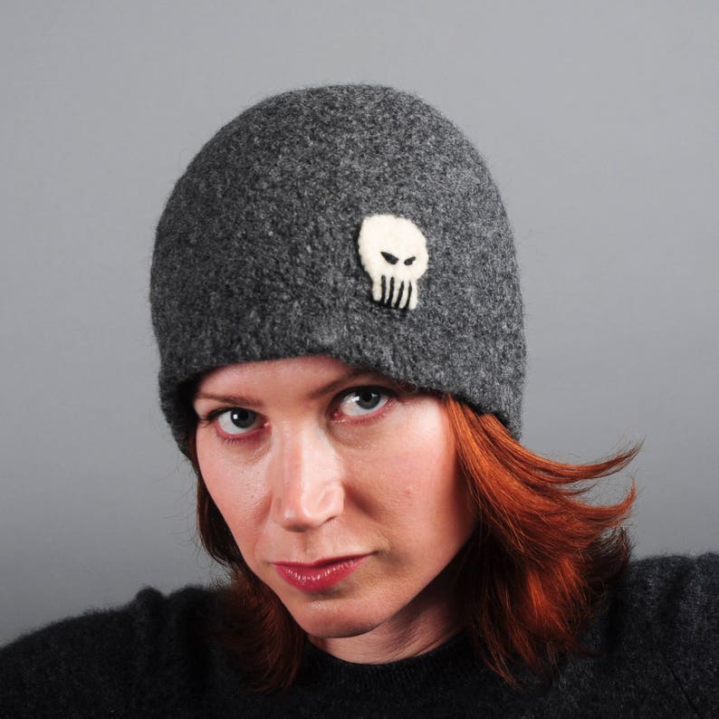 May include: A gray wool beanie with a white felt skull design. The beanie is worn by a person with red hair.