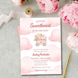 Puede incluir: Una invitación para baby shower con un diseño rosa y blanco. El texto dice "A Little Sweetheart Is On The Way!" con una ilustración de un osito de peluche en un vagón. Incluye detalles como la fecha, hora y lugar del evento.