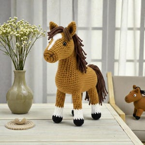 Può includere: Statuetta di cavallo all'uncinetto nei toni del marrone, con una macchia bianca sul muso e una criniera e coda marrone scuro. Il cavallo ha calzini bianchi e zoccoli neri. È visibile anche una versione più piccola del cavallo.