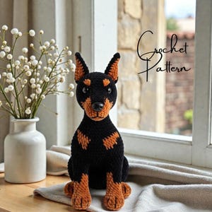 May include: A crocheted Doberman Pinscher dog in black and tan, sitting on a neutral-colored surface. The dog has a black body with tan markings on its chest, legs, and ears. The text "Crochet Pattern" is written in cursive.