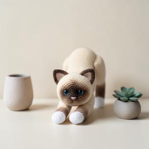 Puede incluir: Un gato siamés tejido a ganchillo en posición de estiramiento. El gato tiene un cuerpo color crema con marcas marrones en la cara, las orejas y la cola. Tiene ojos azules y patas blancas. Una pequeña planta suculenta en una maceta gris y un jarrón de cerámica están en el fondo.