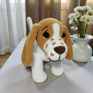 Könnte beinhalten: Handgefertigtes Häkelhundespielzeug in Weiß, Braun und Beige. Der Hund hat große schwarze Augen, eine braune Nase und Schlappohren. Das Spielzeug steht auf einer grauen Oberfläche in der Nähe einer Vase mit weißen Blumen.
