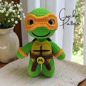May include: A handmade green crochet turtle doll with an orange mask, brown belt, and yellow shell. The doll has black eyes and a smiling face. The text "Crochet Pattern" is written in cursive on the right side.