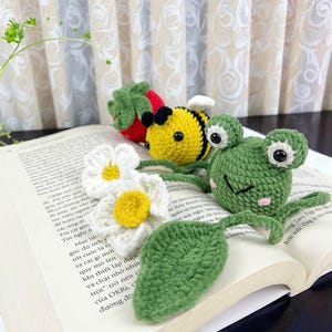 May include: A collection of handmade crochet items, including a red strawberry, a yellow and black bee, a white daisy with a yellow center, a green leaf, and a green frog with large eyes. The items are placed on an open book.