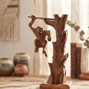 May include: A carved wooden sculpture featuring a monkey hanging from a branch attached to a tree trunk base. The monkey has light-colored circular eyes and a textured body, while the tree trunk displays natural wood grain patterns and knots.