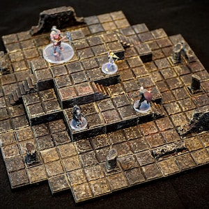 May include: A detailed, multi-tiered dungeon terrain set with a textured, stone-like appearance. Miniature figures are placed on the set, suggesting a tabletop role-playing game. The set features stairs, pillars, and varying levels.