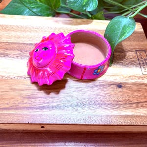 May include: A pink, hand-painted ceramic trinket box with a lion face on the lid. The box is open, revealing a light brown interior. The box sits on a wooden cutting board. The lid has a sun-like design. The box is decorated with blue flowers.