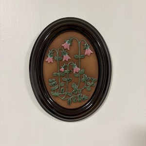 May include: An oval wall plaque featuring a raised ceramic relief of pink bell-shaped flowers with green stems and leaves set against a tan background. The piece is encased in a dark brown glossy frame with a smooth, rounded edge profile.