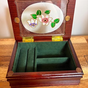May include: A rectangular wooden jewelry box with a dark mahogany finish and a hinged lid. The lid features an oval glass insert decorated with a painted floral design of white and purple flowers with green leaves. The interior is lined in dark green velvet.